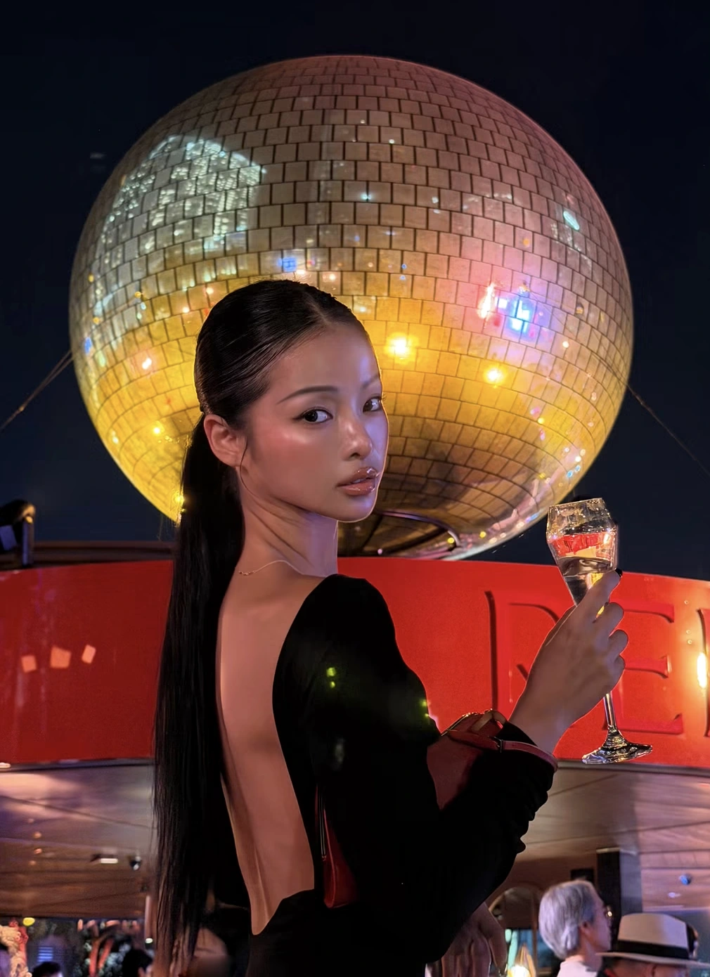 A guest in a black evening dress raises a glass beneath the poolside disco ball