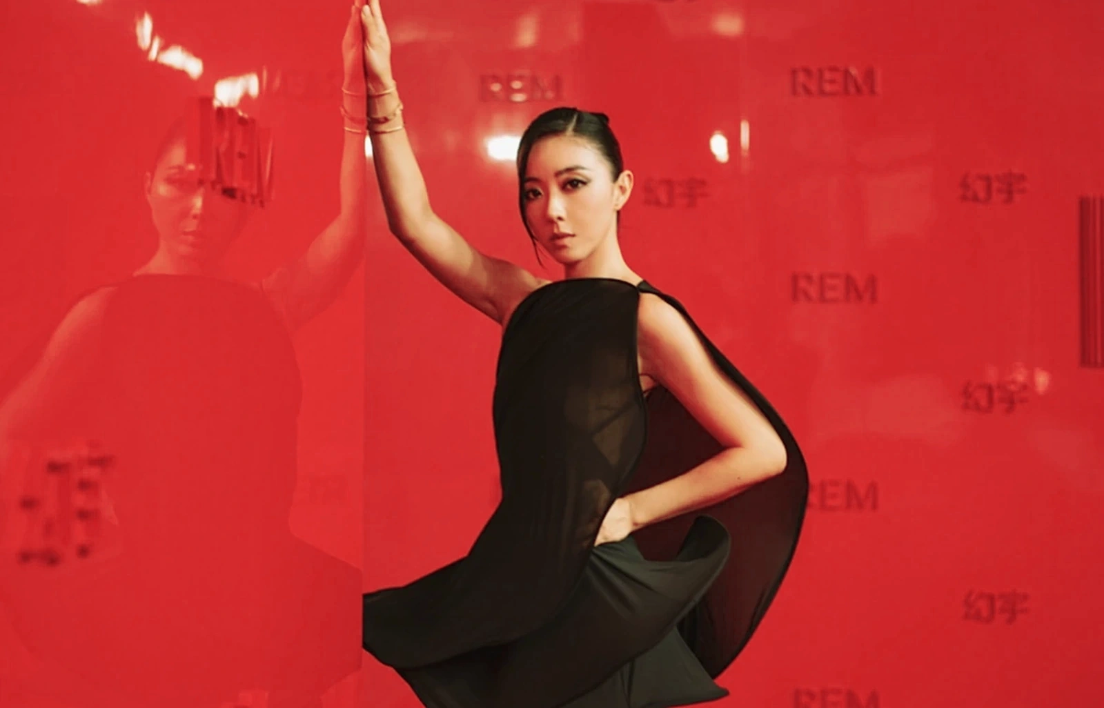 A black sculptural dress reflected in REM’s glossy red carpet backdrop
